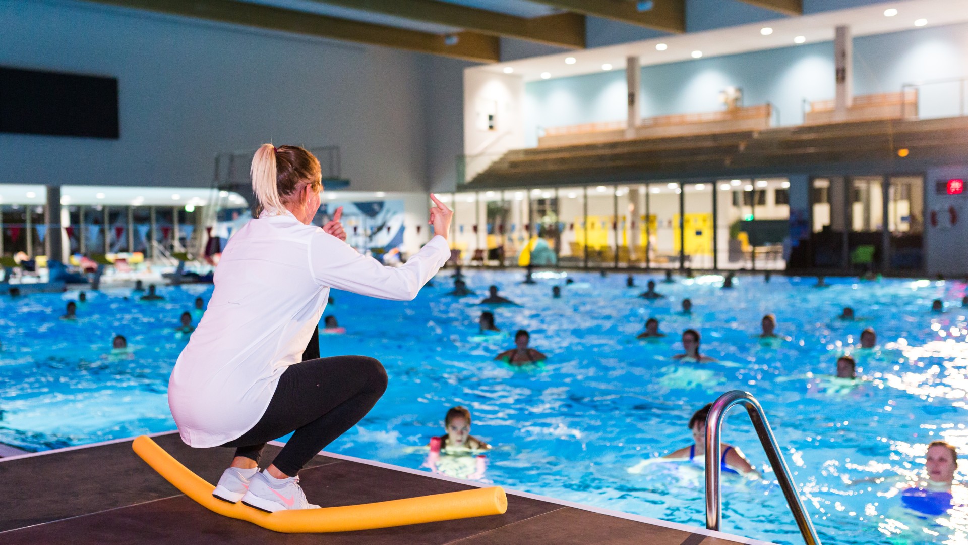 Aquafitness-Trainerin hockend auf einer Poolnudel auf der Bühne erklärt den Teilnehmer eine Übung, © SekundenstillFotografie