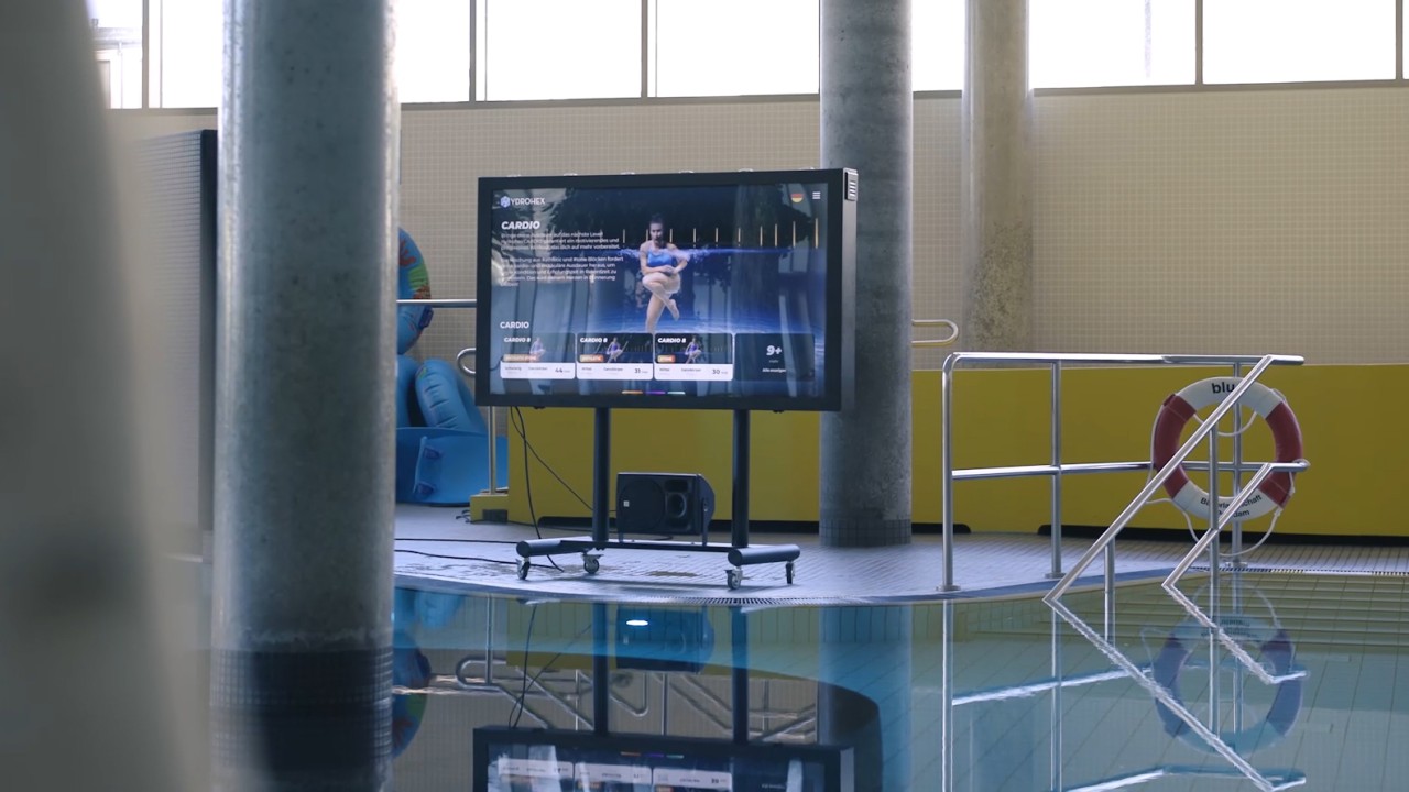 Hydrohex Aquafitness im blu: Auf dem Bildschirm am Beckenrand beginnt eine virtuelle sportliche Frau mit dem Warm-UP im Wasser