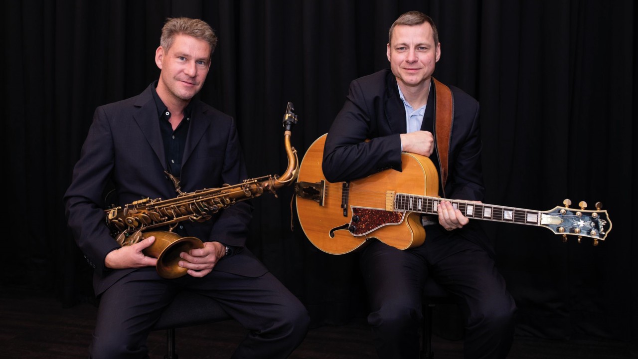 Sebastian Hillmann am Saxophon und Michael Hadrisch an der Gitarre sitzend vor einem schwarzen Vorhang
