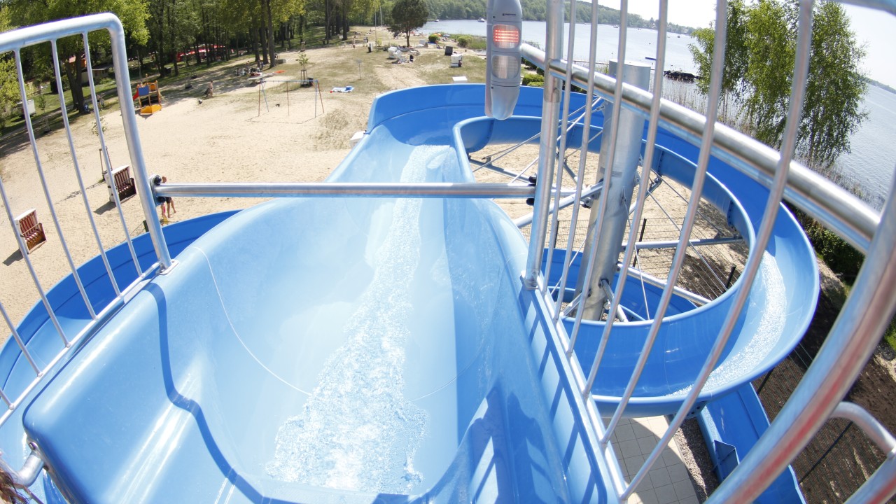 Blaue Wasserrutsche im Waldbad Templin mit Blick auf Sandstrand, Badesee und Bäume; Wasser fließt durch die Rutsche