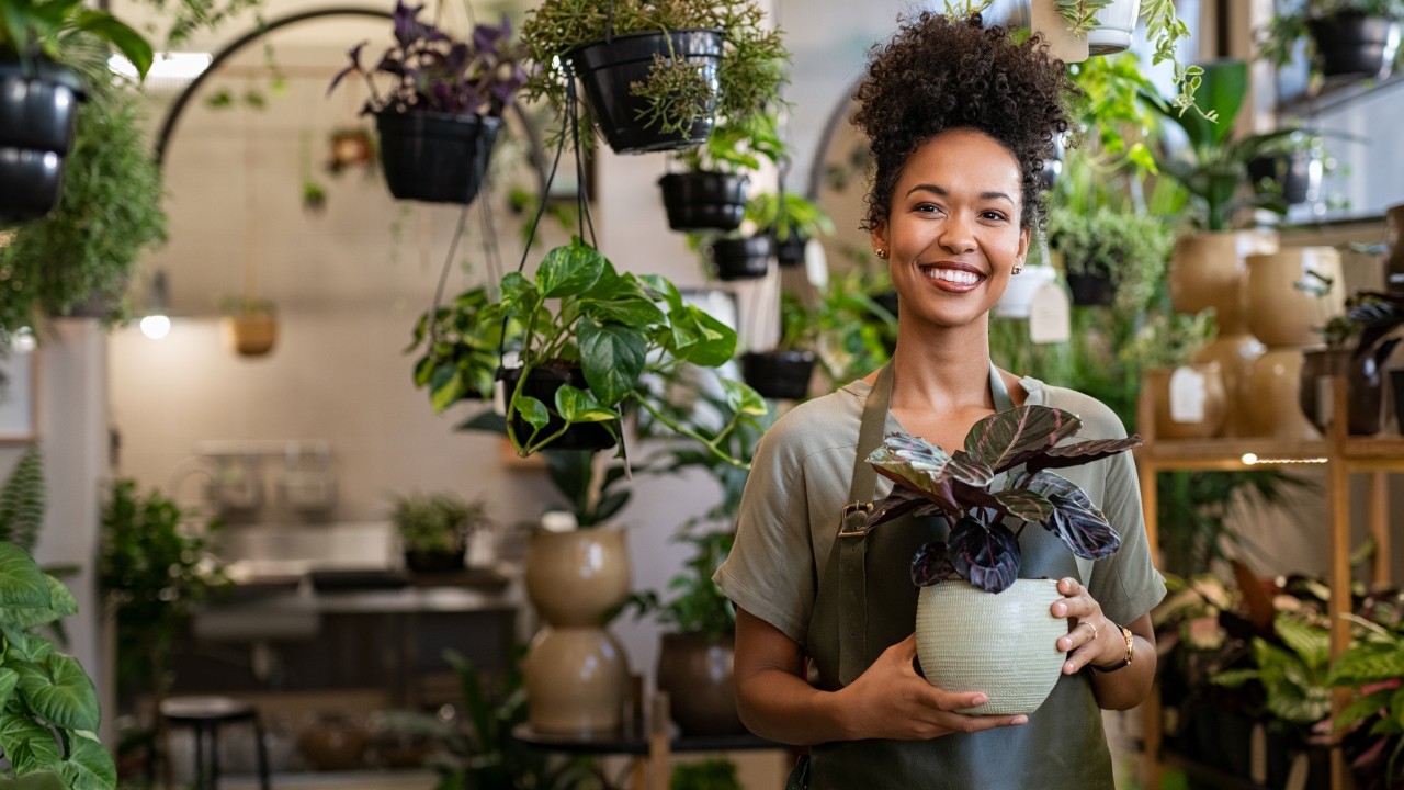 Kleinunternehmerin mit einer Pflanze in den Händen lächelt in ihrem Blumenladen, © Rido – stock.adobe.com