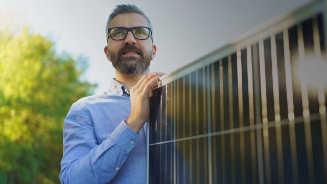 Geschäftsmann steht im Garten und hält ein Solarmodul in den Händen, © Halfpoint – stock.adobe.com