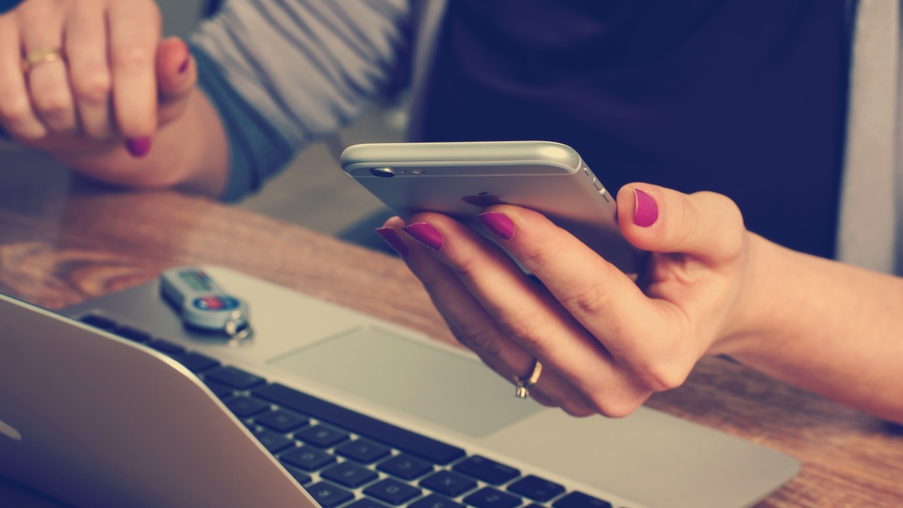 Frau sitzt am Schreibtisch vor dem Laptop und hält ihr Smartphone in der Hand