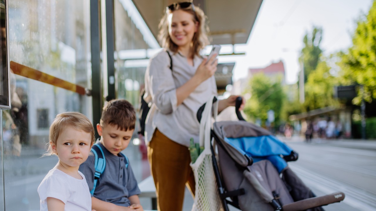 Frau mit zwei Kindern und Kinderwagen warten an Haltestelle